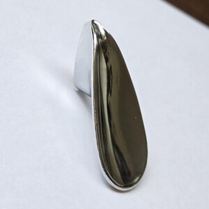 Peut inclure: Un bouton de meuble en chrome poli avec un design lisse et allongé en forme de goutte. La surface métallique est hautement réfléchissante, présentant une silhouette incurvée et effilée qui s'affine vers le haut et s'élargit à la base.