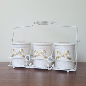 May include: Three white ceramic canisters with lids, gold bow details, and a crackle effect, held in a white metal rack with a handle. Ideal for kitchen storage, each canister is approximately 10 cm tall.