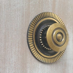 Peut inclure: Gros plan sur un bouton de meuble décoratif. Le bouton présente un design circulaire avec une finition dorée et une base noire. L'anneau extérieur a un motif strié et le centre a un petit cercle surélevé.
