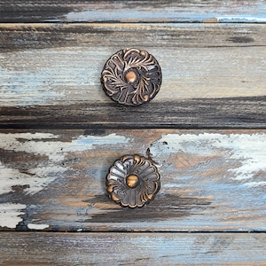 May include: Two ornate, round, copper-colored drawer pulls with a floral design. The pulls are mounted on a distressed wooden surface.