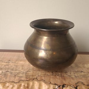 May include: A vintage brass pot with a rounded body and a wide, slightly flared rim. The pot has horizontal bands around its circumference and a warm, golden-brown colour. It sits on a wooden surface with a unique, natural grain pattern.