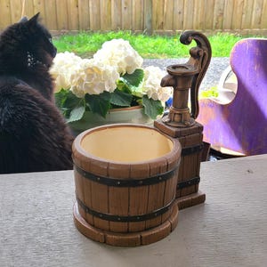May include: A decorative brown planter with a water pump design. The planter has a barrel shape with metal bands. The pump is a dark brown colour. The interior of the planter is a light beige colour.