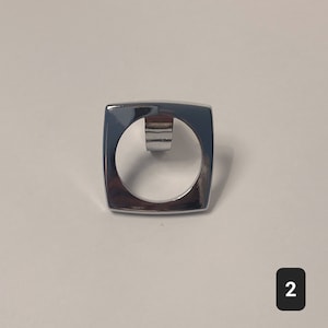 May include: A chrome-plated metal ring pull with a square frame and a circular opening. The ring pull is designed for opening a container or lid.