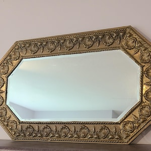 Peut inclure: Miroir octogonal avec un cadre doré orné. Le cadre présente un motif répété de coquillages et de cordes. Le miroir est rectangulaire avec des bords biseautés. Un lézard décoratif et une œuvre d'art encadrée sont visibles en arrière-plan.