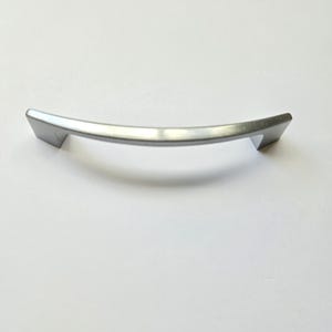 May include: A silver-toned metal cabinet handle with a smooth, arched, and slightly curved design. The handle features two angled mounting points at each end, providing a sleek and functional grip for drawers or cabinet doors in a home setting.