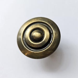 May include: A circular cabinet knob with an antique brass finish. The knob features a raised central dome surrounded by two concentric circular grooves, creating a stepped profile. The metal surface has a weathered, dark gold appearance.