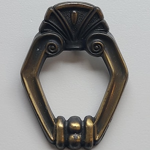 May include: Antique brass-toned drawer pull with an ornate design. The pull features a decorative top with a fan-like pattern and a hexagonal handle. The handle has a series of vertical bars and a central rounded detail. A vintage hardware piece.