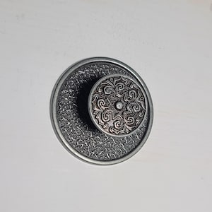 May include: A round metal cabinet knob featuring a dark, textured base plate and a smaller, raised circular center piece. The center piece displays an intricate, embossed scrollwork pattern in a metallic finish, contrasting with the rough, dark surface of the larger circular back plate.