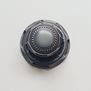 May include: A detailed, antique-style metal knob with a circular design. The knob features a central, raised circle surrounded by a rope-like texture, and multiple concentric rings. The color is a muted, dark gray.