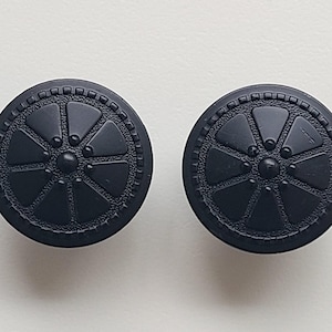 May include: Two black, round cabinet knobs. Each knob features a central circle with a series of triangular shapes radiating outwards, surrounded by a decorative border. The knobs are made of a matte material.