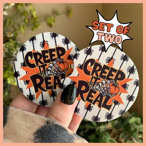 May include: Set of two white round coasters with a black spider web pattern and the text "Creep It Real" in black and orange letters. The coasters are in a hand.
