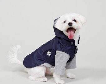 May include: A white dog wearing a navy blue hooded raincoat with a grey shirt underneath. The dog is sitting and looking at the camera with its tongue out.