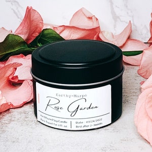 May include: A black tin candle with a white label that reads "Earthy-Marpe Rose Garden Hand-Poured Soy Candle Wax Transit 4 fl oz Date 01/24/2022 Best after 2 weeks" The candle is surrounded by pink roses.