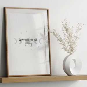 May include: A framed print with the text "Anything for our Moon" and a crescent moon design in gray. The print is on a wooden shelf with a white vase and dried white flowers.