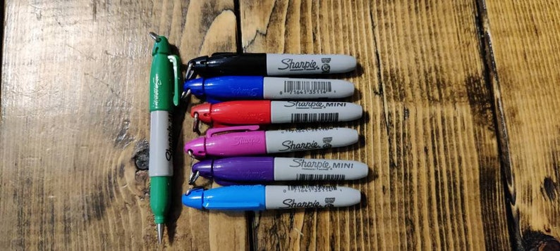 May include: A set of eight Sharpie brand permanent markers in various colors, including black, blue, red, pink, purple, and light blue. The markers are all mini size and have a silver cap with the Sharpie logo.