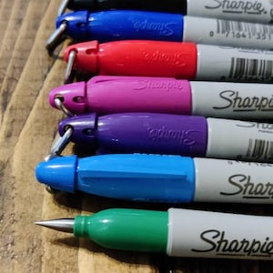 May include: A set of six Sharpie permanent markers with different colored caps and ink. The markers are arranged in a row from top to bottom, with the black marker at the top and the green marker at the bottom. The markers are all labeled "Sharpie" and have a barcode on the side.