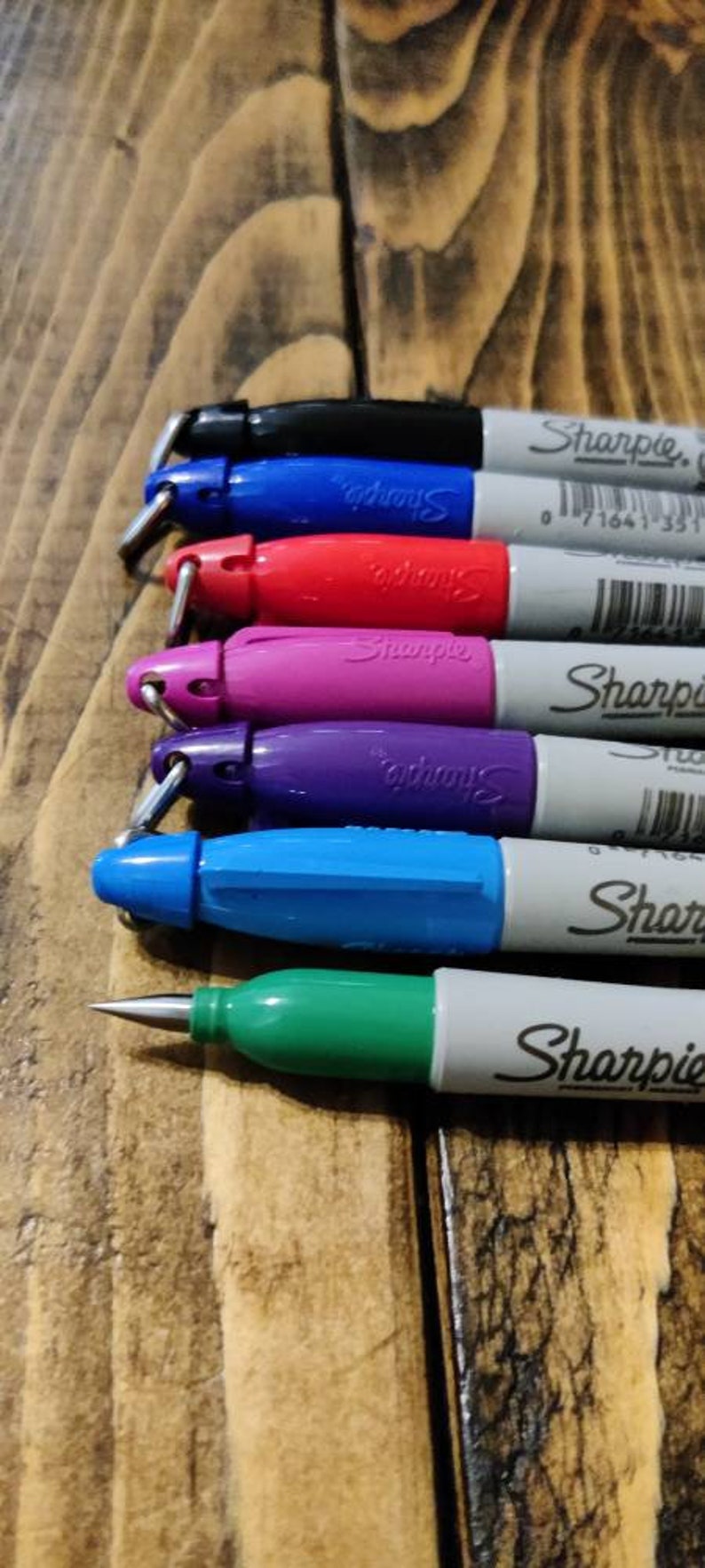 May include: A set of six Sharpie permanent markers with different colored caps and ink. The markers are arranged in a row from top to bottom, with the black marker at the top and the green marker at the bottom. The markers are all labeled "Sharpie" and have a barcode on the side.