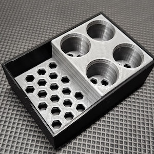 Puede incluir: Un organizador de dos tonos con una bandeja exterior negra y una inserción plateada. La inserción presenta un patrón de panal y cuatro compartimentos circulares. El organizador está diseñado para guardar objetos pequeños.