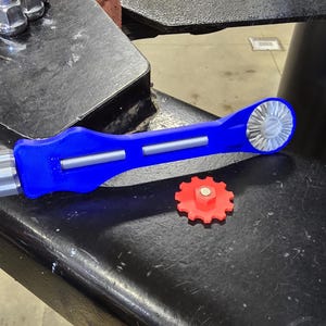 May include: A blue and silver tool with a red gear. The tool has a handle and a gear on one end. The gear is designed to fit into a larger gear.