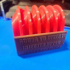 K&ouml;nnte beinhalten: Ein 3D-gedruckter Halter f&uuml;r Z&uuml;ndkerzen, beschriftet mit "ACURA V6 DIRECT INJECTED UPPER INTAKE PORT PLUGS". Der Halter ist braun und enth&auml;lt sechs rote Z&uuml;ndkerzen.