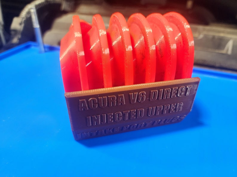 K&ouml;nnte beinhalten: Ein 3D-gedruckter Halter f&uuml;r Z&uuml;ndkerzen, beschriftet mit "ACURA V6 DIRECT INJECTED UPPER INTAKE PORT PLUGS". Der Halter ist braun und enth&auml;lt sechs rote Z&uuml;ndkerzen.