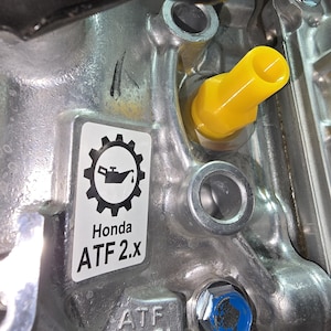 May include: Close-up of a metallic engine component with a yellow cap and a blue bolt. A sticker displays a gear and oil can icon, with the text "Honda ATF 2.x" and "ATF LEVEL".