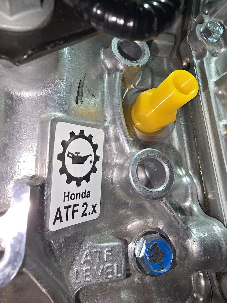 May include: Close-up of a metallic engine component with a yellow cap and a blue bolt. A sticker displays a gear and oil can icon, with the text "Honda ATF 2.x" and "ATF LEVEL".