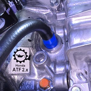 May include: Close-up of a metallic engine component with a black hose and blue fitting. A sticker reads "Honda ATF 2.x" with a gear and oil can graphic. An orange bolt is visible.