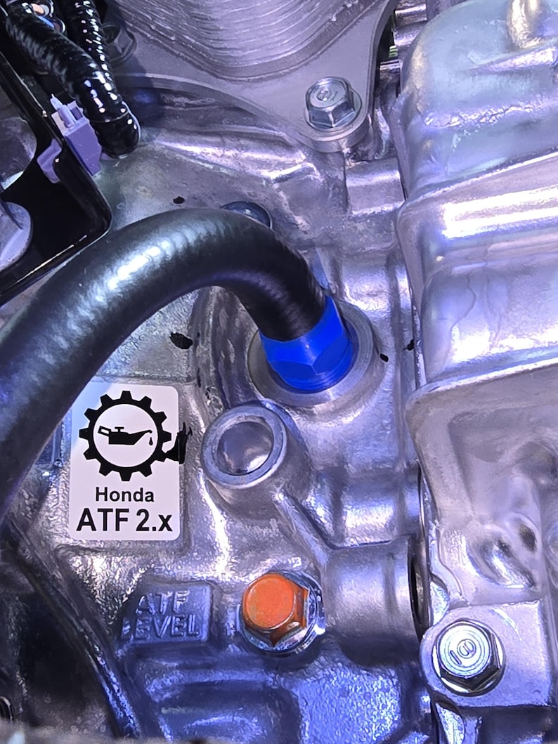 May include: Close-up of a metallic engine component with a black hose and blue fitting. A sticker reads "Honda ATF 2.x" with a gear and oil can graphic. An orange bolt is visible.