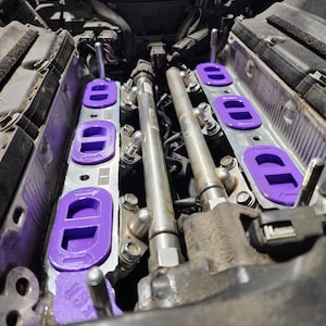 May include: Close-up of a car engine with purple intake manifold spacers. The spacers are designed to improve airflow and performance.