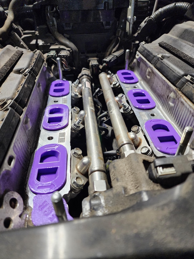 May include: Close-up of a car engine with purple intake manifold spacers. The spacers are designed to improve airflow and performance.
