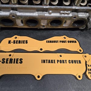 May include: An overhead shot displays three tan K-Series engine port covers with black text. The covers are labeled "Exhaust Port Cover", "Intake Port Cover", and "Coolant Passage Cover". The text "Do Not Place On A Hot Engine" is also visible.