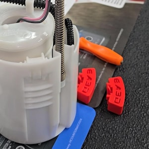 May include: A white automotive fuel pump assembly with a spring, black wire, and red clips. An orange tool and a blue card with white text are also visible. The background is a dark surface.