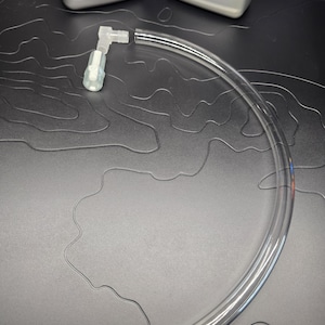 May include: A clear, curved plastic tube with a white plastic connector. Two gray bottles with black labels are in the background. The labels read "HONDA" and "1 U.S. QT/946 ML". The items are on a black surface.
