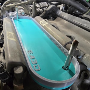 May include: A teal and gray 3D-printed automotive part installed in an engine compartment. The part has the text "E43D" and "HEAT RESISTANT TO 230". The part is designed to fit around two bolts.