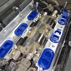 May include: An automotive engine component featuring a metallic silver intake manifold base with several bright blue oval-shaped gaskets installed. Two black cylindrical ignition coils are positioned centrally, secured by metal bolts and hardware.