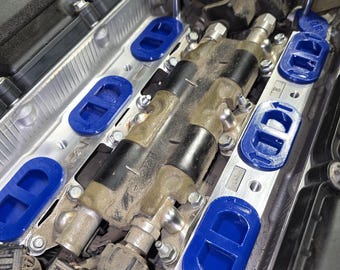 May include: An automotive engine component featuring a metallic silver intake manifold base with several bright blue oval-shaped gaskets installed. Two black cylindrical ignition coils are positioned centrally, secured by metal bolts and hardware.