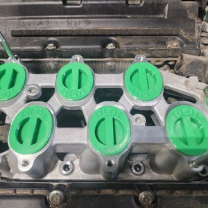 K&ouml;nnte beinhalten: Nahaufnahme eines Automotors mit sechs gr&uuml;nen Ventildeckelkappen. Die Kappen sind oval geformt mit einem erh&ouml;hten Grat in der Mitte. Der Motor ist aus Metall und hat eine silberne Oberfl&auml;che.