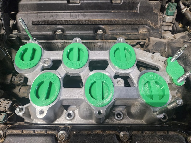 K&ouml;nnte beinhalten: Nahaufnahme eines Automotors mit sechs gr&uuml;nen Ventildeckelkappen. Die Kappen sind oval geformt mit einem erh&ouml;hten Grat in der Mitte. Der Motor ist aus Metall und hat eine silberne Oberfl&auml;che.