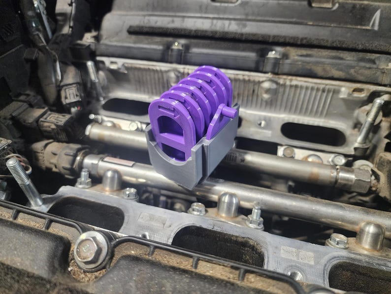 May include: A close-up of a purple and gray engine part with a series of circular openings. The part is sitting on top of a car engine.