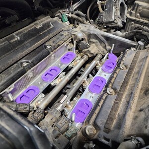 May include: Close-up of a car engine with purple intake manifold spacers. The spacers are installed on the intake manifold and are connected to the fuel injectors.