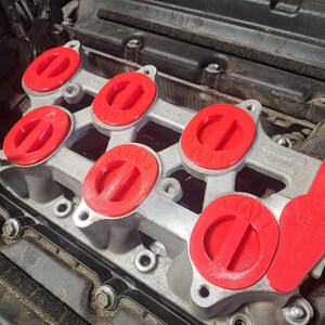 K&ouml;nnte beinhalten: Nahaufnahme eines Automotors mit sechs roten Ansaugkr&uuml;mmerabdeckungen. Die Abdeckungen haben ein erhabenes "100"-Logo.