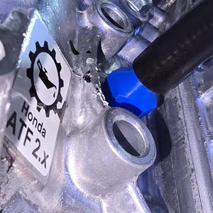 May include: Close-up of a silver metal engine component with a blue cap and a black hose. A sticker with the Honda logo and the text "ATF 2.X" is visible.