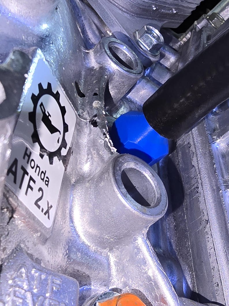 May include: Close-up of a silver metal engine component with a blue cap and a black hose. A sticker with the Honda logo and the text "ATF 2.X" is visible.