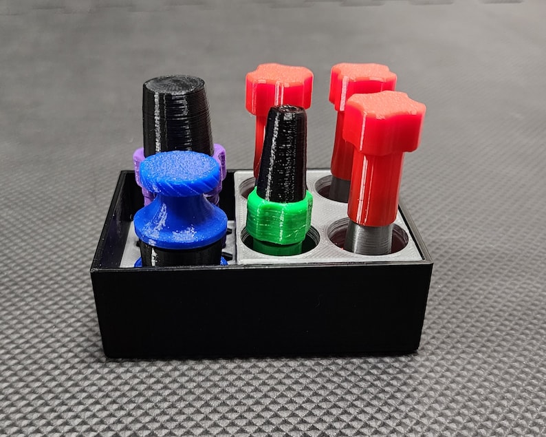 Peut inclure: Un organisateur rectangulaire noir contient plusieurs objets color&eacute;s imprim&eacute;s en 3D. Ceux-ci comprennent un objet bleu en forme de champignon, une pi&egrave;ce cylindrique noire et des objets rouges en forme d'&eacute;toile. L'organisateur est sur une surface textur&eacute;e.