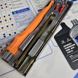May include: A gray plastic tool with orange handles for filling automatic transmission fluid. The tool is labeled "EPS RECALL" and is sitting on a white surface with a chart of Honda and Acura vehicles and their corresponding fluid types. The chart includes the text "Automatic Transmission Fluid BEVF - Type PZ".