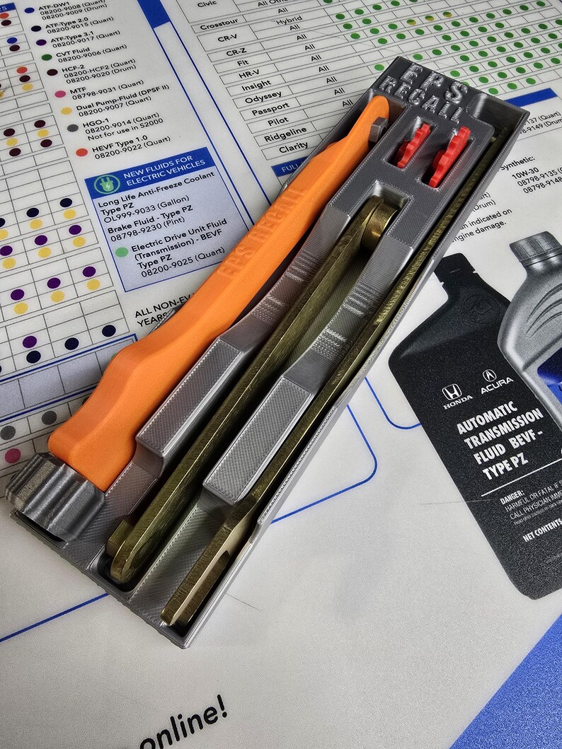 May include: A gray plastic tool with orange handles for filling automatic transmission fluid. The tool is labeled "EPS RECALL" and is sitting on a white surface with a chart of Honda and Acura vehicles and their corresponding fluid types. The chart includes the text "Automatic Transmission Fluid BEVF - Type PZ".