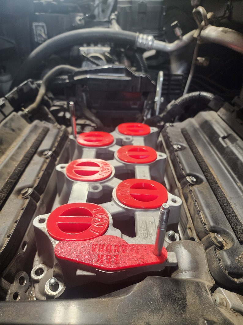 K&ouml;nnte beinhalten: Nahaufnahme eines Automotors mit roten Plastikabdeckungen &uuml;ber den Z&uuml;ndkerzenl&ouml;chern. Die Abdeckungen tragen den Text "EGR" und "AURA".