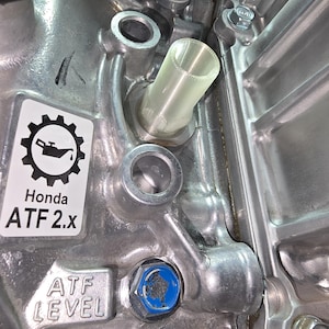May include: Close-up of a metallic engine component with a clear plastic tube inserted. The image features a sticker with a gear and oil can icon, and the text "Honda ATF 2.x". A blue bolt is visible, along with the text "ATF LEVEL".