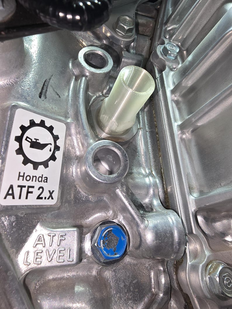May include: Close-up of a metallic engine component with a clear plastic tube inserted. The image features a sticker with a gear and oil can icon, and the text "Honda ATF 2.x". A blue bolt is visible, along with the text "ATF LEVEL".